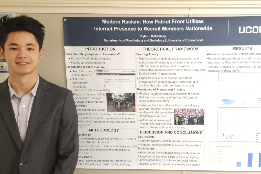 Kyle L. Makalusky stands in front of a research poster entitled "Modern Racism: How Patriot Front Utilizes Internet Presence to Recruit Members Nationwide"