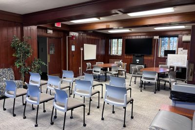 Lounge with two small conference tables, rows of rolling chairs, lounge seating, and a wall-mounted TV.