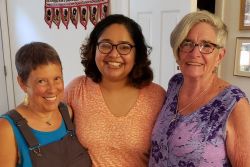 Three people - one in overalls and a blue shirt, another in an orange shirt, and the third in a purple dress, pose arm-in-arm for a photograph.